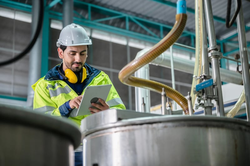 Técnico industrial analisando backlog de manutenção em tablet dentro de fábrica com equipamentos ao fundo