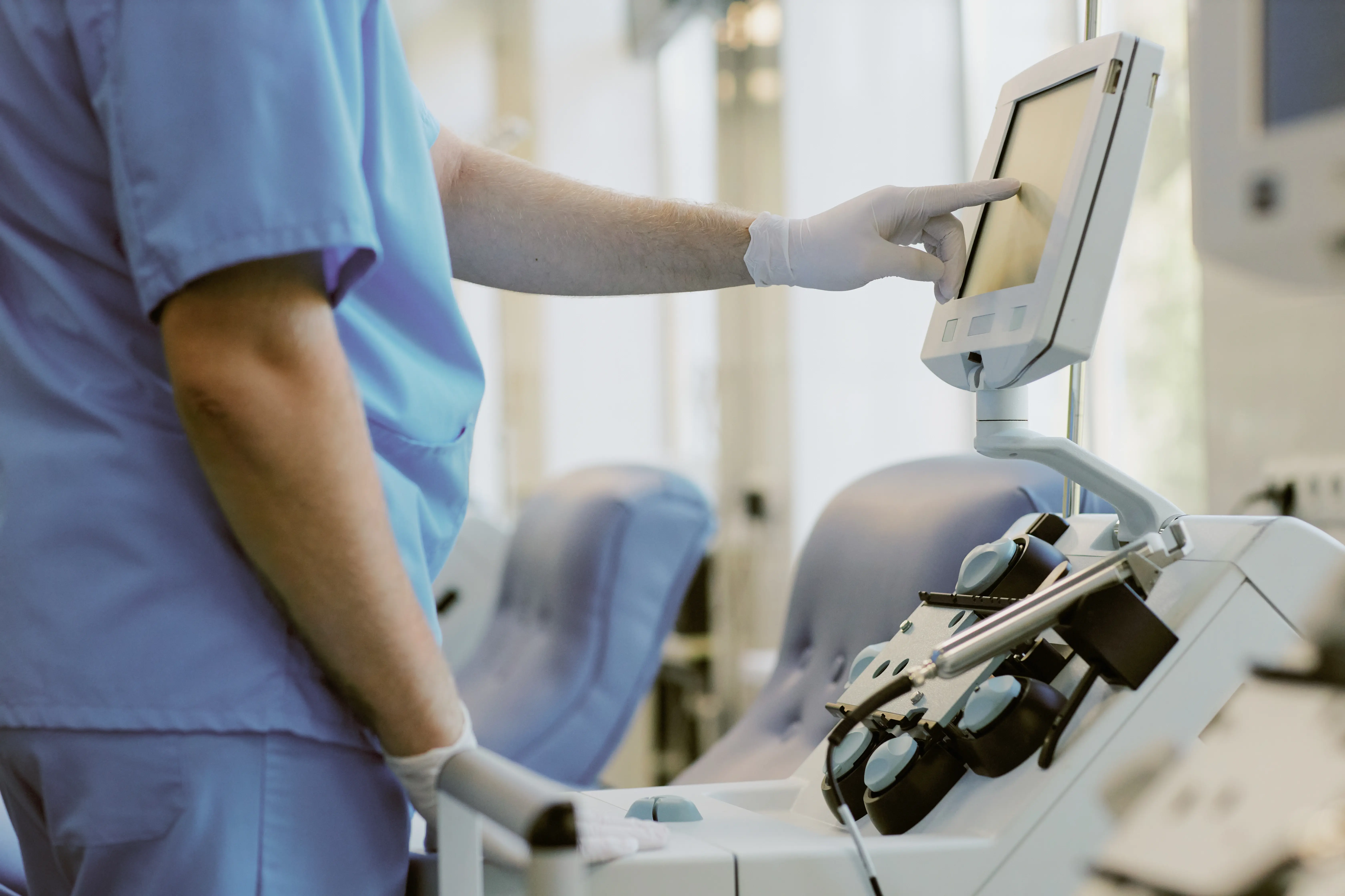 Profissional de saúde usando luvas e uniforme azul ajusta a tela sensível ao toque de um equipamento médico em uma sala clínica.